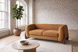 Greccio boucle loveseat in caramel in a white-brick loft with arched windows, a fig tree and a round travertine table