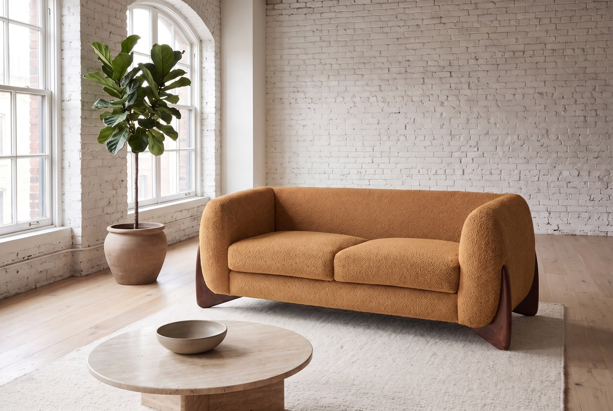 Greccio boucle loveseat in caramel in a white-brick loft with arched windows, a fig tree and a round travertine table