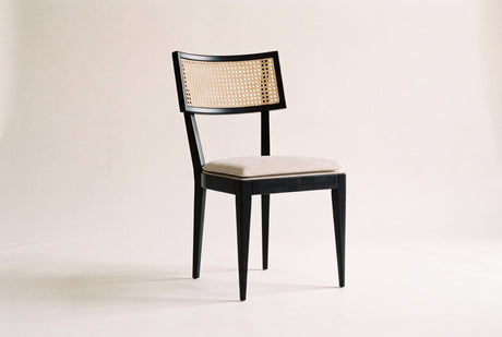 Harper dining chair with a black oak frame, natural woven cane back and oatmeal cushion, three-quarter view on a warm off-white studio backdrop