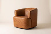 Hollis swivel chair in Saddle leather, three-quarter view on a warm off-white studio backdrop: a rounded barrel back curving