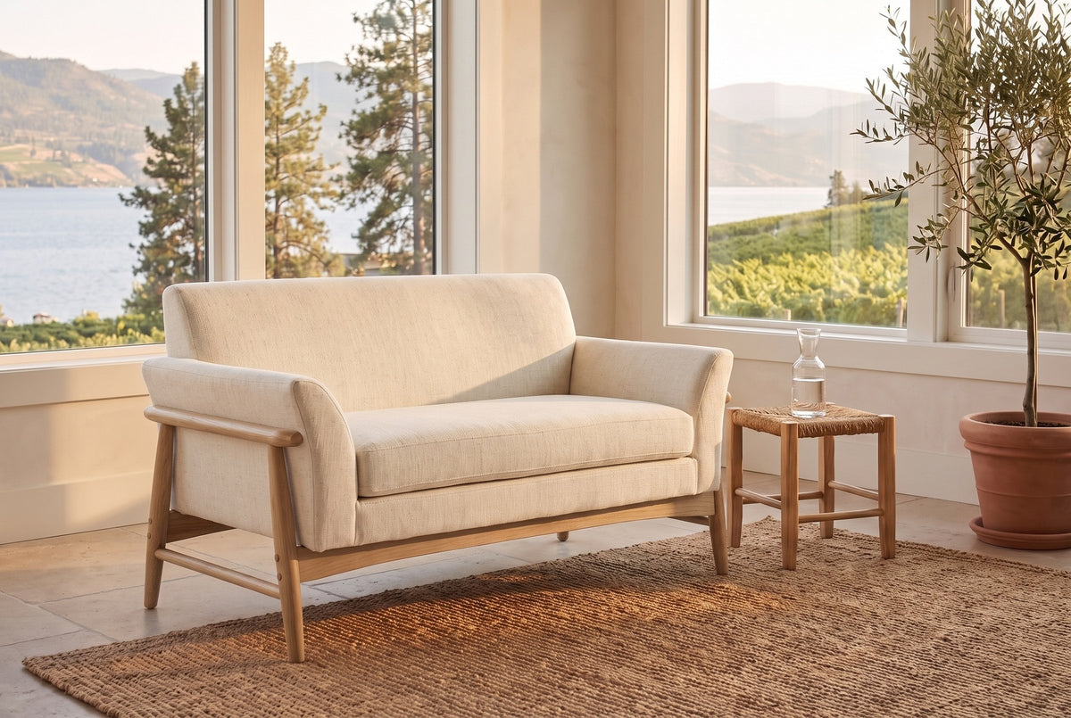 Jesi loveseat in beige woven fabric on a natural-finish frame in a Kelowna lake house sunroom with a jute rug, a rush stool and a view of the lake and vineyard hills
