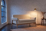 Jesi loveseat in beige woven fabric on a natural-finish frame against a whitewashed brick wall in a Montreal loft at dusk, a brass floor lamp lit beside it