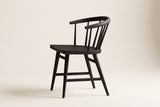 Side view of one black oak Lauryn dining chair on an off-white backdrop, with the low curved back rail, the slim spindles, the carved seat and the four splayed legs with their stretchers