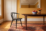 One black oak Lauryn dining chair with a curved spindle back at the end of an oval walnut table with a blue and white bowl of red apples and a copper jug of orange marigolds, in an ochre yellow room with a white panelled door and white mouldings, on an oak parquet floor with a red and blue patterned rug in warm evening light