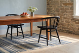 Two black oak Lauryn dining chairs with curved spindle backs at a long cherry wood table with a speckled stoneware bowl of peaches and a grey crock of white Queen Anne’s lace, beside an exposed limestone wall and a pale blue plaster wall, on pale pine floorboards with a blue and cream striped rug in morning sun