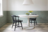 Two black oak Lauryn dining chairs with curved spindle backs at a round white pedestal table with a celadon bowl of green figs and a white vase of yellow tulips, under a white pleated pendant, by a celadon green tiled wall on a cream and grey checkerboard stone floor