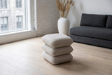 A Lavagna ottoman in Beige on a pale oak floor in a whitewashed brick loft in Old Montreal, a charcoal wool sofa behind