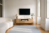 The Livia media console in Natural below a switched-off TV, in a tall Lisbon apartment with plaster cornices, pine floorboards, a blue and white rug and a curved ivory sofa