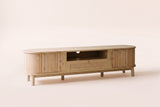 The Livia media console in Natural, front three-quarter view: pale wood with rounded ends, curved reeded doors and a rattan drawer, on an off-white studio background