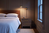 Lodi nightstand in Brown Oak beside a bed in a taupe Ottawa townhouse bedroom at dusk, a brass wall lamp lit above it