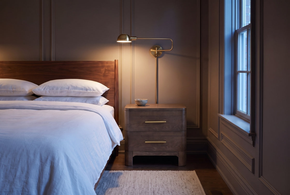 Lodi nightstand in Brown Oak beside a bed in a taupe Ottawa townhouse bedroom at dusk, a brass wall lamp lit above it
