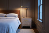 Lodi nightstand in Natural beside a bed in a taupe Ottawa townhouse bedroom at dusk, a brass wall lamp lit above it