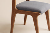 Close-up of a Lucrezia dining chair in the Walnut finish: grey fabric seat cushion inset in the oak seat frame and tapered legs, on an off-white background