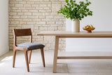 One Lucrezia dining chair in the Walnut finish with a grey fabric seat, pulled up at the end of a long pale oak trestle table in a front three-quarter view, with green linden branches in a cream vase and a wooden bowl of yellow pears, beside a limestone fieldstone wall on pale stone floors