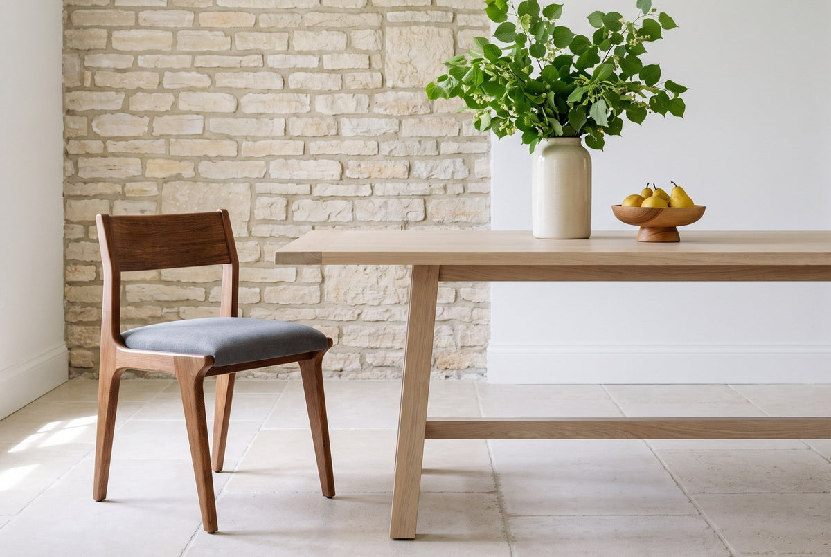 One Lucrezia dining chair in the Walnut finish with a grey fabric seat, pulled up at the end of a long pale oak trestle table in a front three-quarter view, with green linden branches in a cream vase and a wooden bowl of yellow pears, beside a limestone fieldstone wall on pale stone floors