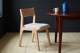 One Lucrezia dining chair in the White Oak finish with an off-white fabric seat, in a three-quarter view and turned toward a round mahogany table with a blue enamel coffee pot and a cream mug, against charcoal shiplap walls on pale painted floors