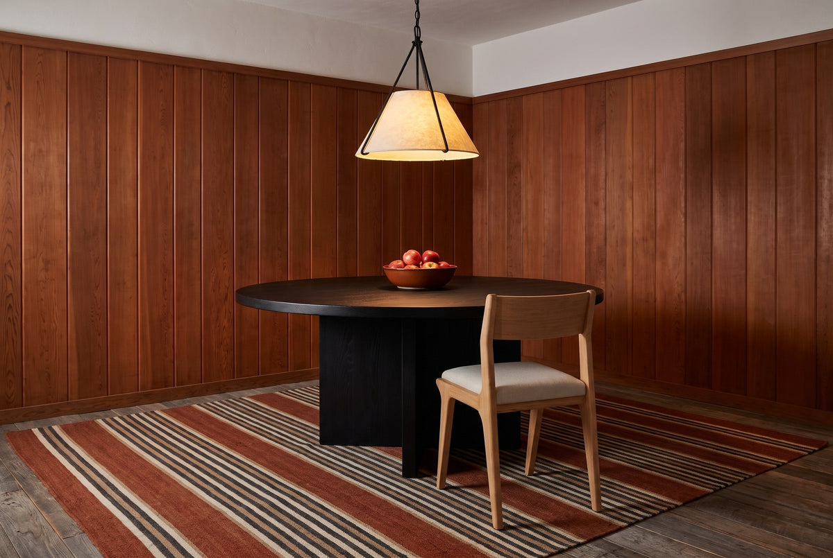 One Lucrezia dining chair in the White Oak finish with an off-white fabric seat, seen from behind and pushed in at a round black oak pedestal table with a bowl of red apples, under a parchment pendant in a cedar-panelled room with a striped wool rug
