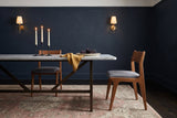 Two Lucrezia dining chairs in the Walnut finish with grey fabric seats at a long white marble table: one tucked in on the far side, the other pulled out past the table end in a side three-quarter view, with three lit taper candles, a mustard linen napkin and a platter of grapes, in a navy room with brass wall sconces and a rose antique rug