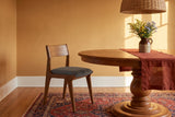 One Lucrezia dining chair in the Walnut finish with a grey fabric seat, pushed in at a round cherry pedestal table in a side three-quarter view, with a rust-red linen runner and a stoneware jug of goldenrod and blue asters beneath a woven rattan pendant, in an ochre room with a patterned rug in afternoon light