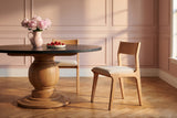 Two Lucrezia dining chairs in the White Oak finish with off-white fabric seats at a round black stone table on a turned oak pedestal: one tucked in on the far side, the other pulled out in the foreground in a side three-quarter view, with pink peonies and strawberries, in a dusty rose panelled room on herringbone oak floors