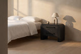Macerata nightstand in black beside a bed in white linen, in a plaster-walled bedroom in late golden light