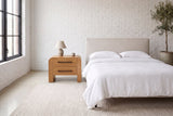 Macerata nightstand in brown beside a bed in white linen, in a white-brick loft bedroom with a tall black-framed window