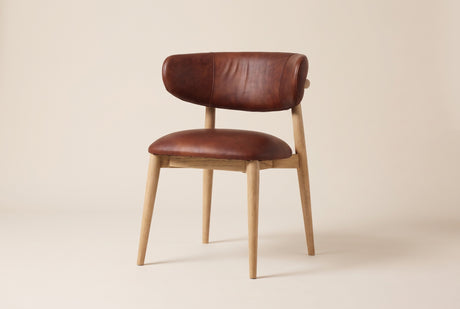 Marlow leather dining chair in Tobacco, three-quarter view on a warm off-white studio backdrop: a deep reddish-brown back and seat on light ash legs