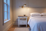 Marsala nightstand in Milk White beside a bed in a panelled Toronto bedroom at dusk, a brass swing-arm lamp lit above it