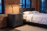 Merano nightstand in Black beside a bed against old brick in a Montreal loft at dusk, a brass wall lamp lit above it