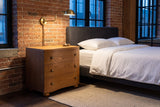 Merano nightstand in Honey Brown beside a bed against old brick in a Montreal loft at dusk, a brass wall lamp lit above it