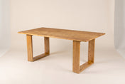 Mireille dining table in Honey, three-quarter view on a warm off-white studio backdrop: a long solid acacia top on two open r