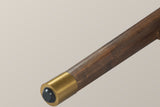 Close-up of one tapered Monteriggioni leg in brushed walnut-finish wood ending in a gold-tone foot cap with a black glide