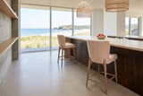 Two Morrovalle counter stools in Champagne Chenille seen from behind along a corten-clad island with a travertine top in a La