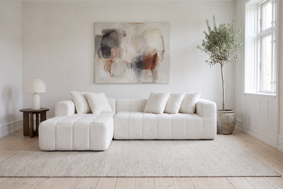 Nembo Boucle Cloud Sectional against a white wall in a Copenhagen apartment living room under an abstract painting, a paper lamp on a dark oak side table by the chaise, an olive tree at the other end and an oatmeal wool rug