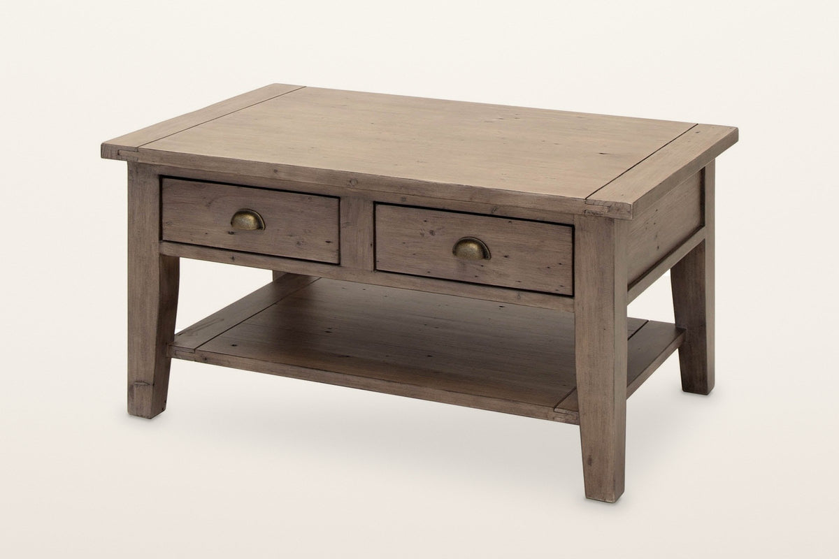 Novella coffee table in Sundried reclaimed pine: two drawers with brass cup pulls and a low open shelf, three-quarter view on a warm white studio backdrop