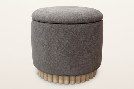 Nyla round storage ottoman in warm charcoal chenille on a reeded light-wood base, seen from slightly above on a warm white studio backdrop