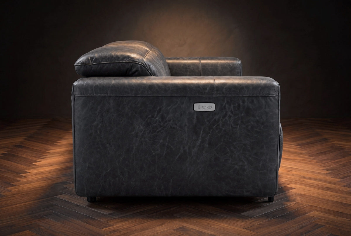 The Octavia loveseat in Aged Slate distressed full leather on a dark herringbone floor, seen from the side, the arm with its control panel and a raised headrest. Shown in Aged Slate, Loveseat.