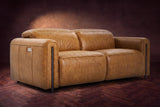 The Octavia loveseat in Antiqued Caramel distressed full leather on a dark herringbone floor, headrests down and footrests closed. Shown in Antiqued Caramel, Loveseat.