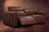 The Octavia loveseat in Burnished Chestnut distressed full leather on a dark herringbone floor, the left recliner open and its headrest raised. Shown in Burnished Chestnut, Loveseat.