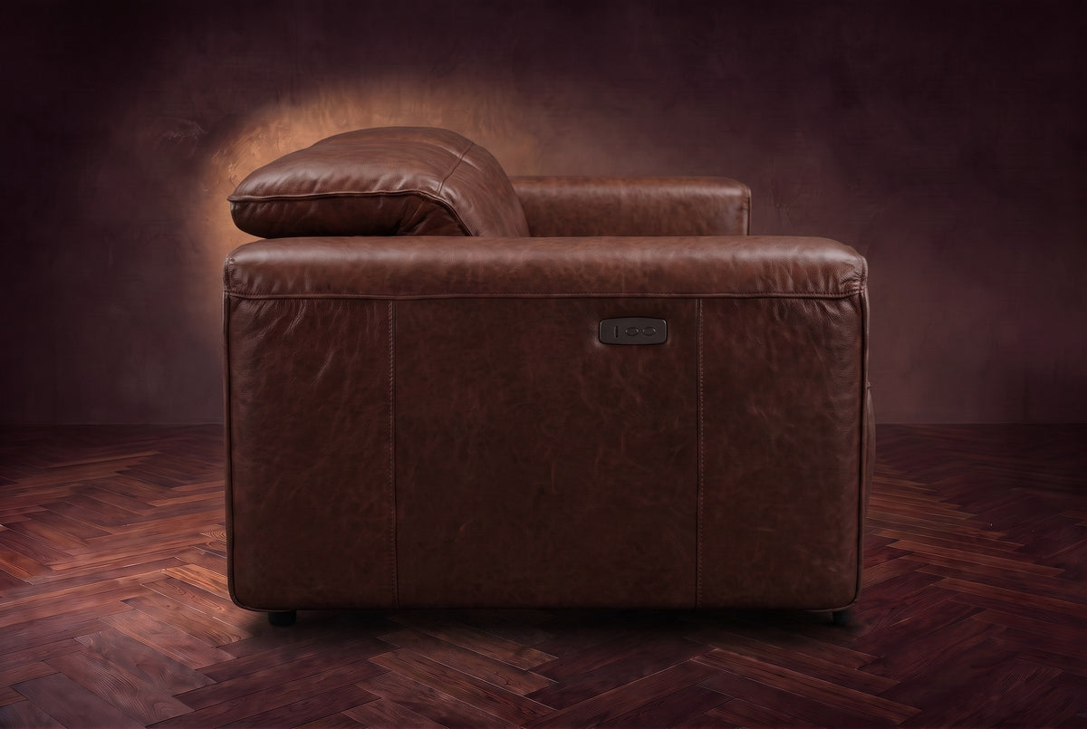 The Octavia loveseat in Burnished Chestnut distressed full leather on a dark herringbone floor, seen from the side, the arm with its control panel and a raised headrest. Shown in Burnished Chestnut, Loveseat.