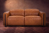 The Octavia loveseat in Tuscan Cognac distressed full leather on a dark herringbone floor, seen from the front, headrests down. Shown in Tuscan Cognac, Loveseat.