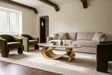 The Ondina coffee table in Gold on an ivory rug between an oat linen sofa and olive velvet armchairs, in a plaster room with dark oak beams and a leaded window
