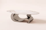 The Ondina coffee table in Gray, front view: a white veined sintered stone oval on one polished gray steel ribbon that curls, dips and scrolls, with a gray sphere resting on it, on an off-white studio background