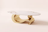 The Ondina coffee table in Gold, front view: a white veined sintered stone oval on one polished gold steel ribbon that curls, dips and scrolls, with a gold sphere resting on it, on an off-white studio background