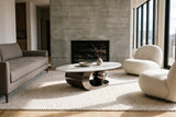 The Oriana coffee table in Gray on a cream rug in front of a grey linen sofa and an ivory lounge chair, beside a board-formed concrete fireplace wall in bright daylight