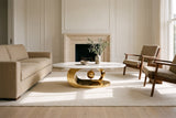 The Oriana coffee table on a cream rug between a sand linen sofa and two walnut lounge chairs, in a white-panelled living room with a pale travertine fireplace and wide oak floorboards