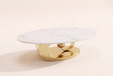 The Oriana coffee table, front three-quarter view: a white veined sintered stone oval top on a gold stainless steel base of a plinth, a ring and a sphere, on an off-white studio background