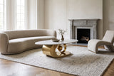 The Oriana coffee table on an ivory wool rug in front of a curved oat boucle sofa, in a tall drawing room with a carved grey stone fireplace and an oak herringbone floor in daylight