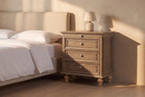Osimo nightstand in Light Brown beside a linen bed in late golden sun, its framed side panel in the light