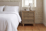Osimo nightstand in Light Brown beside a bed with white linen against a limestone wall, a lamp and a stoneware bowl on top